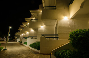 Coastal resort features lit balconies along cobblestone path at night. Warm lighting enhances architectural angles, creating serene atmosphere under dark sky