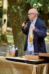Mature bald male with glasses holding amber necklace during outdoor lecture. Standing by table with jars, under trees, speaking into microphone. Bright daylight