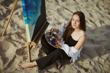Young female artist, seated on sandy beach, immerses herself in painting. Canvas exhibits vibrant...