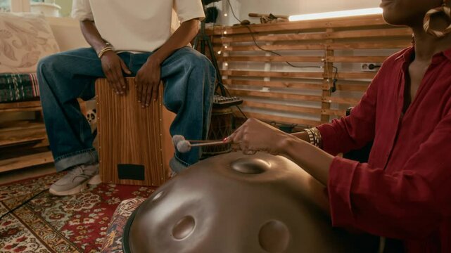 Cropped shot of young African American woman sitting on the floor and beating glucophone with drumsticks while playing with guy sitting on wooden drum during repetition