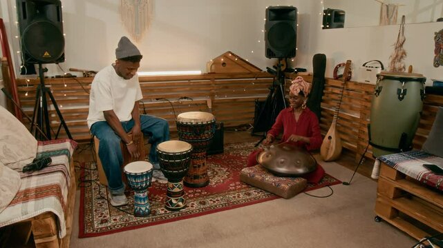 Two young African American musicians playing ethnic drums while girl sitting on the floor, playing glucophone and looking at her boyfriend beating wooden timbal