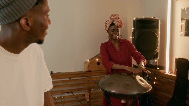 Young smiling African American male and female musicians in casual apparel looking at one another and playing ethnic drums while enjoying musical repetition in home studio