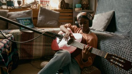 Talented African American girl in headphones sitting on the floor by couch in home studio and playing acoustic guitar in front of microphone during repetition - Powered by Adobe