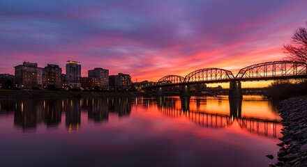 Naklejka premium This picturesque urban landscape features a stunning sunset over a river, showcasing a grand bridge and city lights beautifully reflected on the water