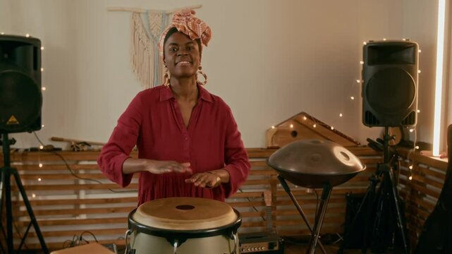 Young cheerful female musician in ethnic accessories and casual dress beating percussion energetically while creating new music and enjoying repetition in home studio