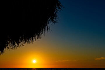 The sun sets on vacation, over the water while sitting on the beach, under an umbrella.