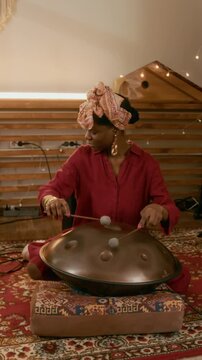 Vertical shot of young African American woman sitting on carpet on the floor of home studio and beating metallic glucophone with drumsticks while creating new ethnic native music