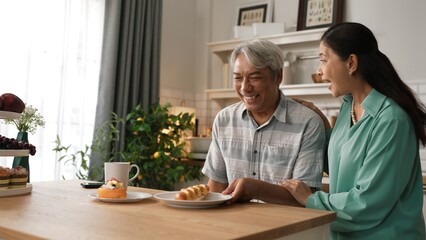 Fototapeta premium Energetic grandmother holding breakfast and serving to her husband while grandfather sitting at modern kitchen. Smiling elder couple spending time together. Retirement lifestyle concept. Myrmidon.