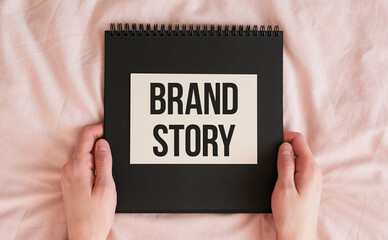 A person is holding a black spiral bound notebook with the word Brand Story written on it.