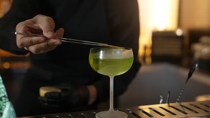 Cropped bartender garnishing caramelized citrus slice on cube ice in green Midori Sour cocktail...