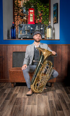 Young jazz musician sitting in an armchair leaning on a tuba resting on the floor, wearing a gray retro-style suit.