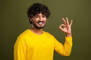 Young man in yellow shirt making an OK hand gesture against green background showing positivity and...