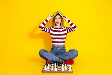 Young woman in striped pullover playfully poses with book on head near chessboard against yellow...