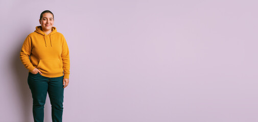 Non-binary person in mustard hoodie stands with hand in pocket against lavender background.