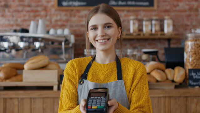 A cheerful young woman in a yellow sweater and apron holds out a card payment terminal in a cozy bakery filled with fresh bread and pastries.
