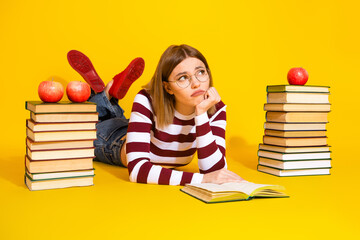 Young woman lying on floor with book and apples, thinking while studying on a vibrant yellow...