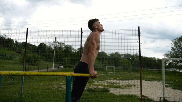 Young sportsman doing push ups on parallel bars at nature. Strong sporty man exercising at sports ground. Male shirtless athlete training hard outdoor. Concept of active sportive lifestyle. Slow mo