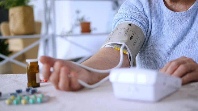 Senior woman using a blood pressure monitor at home, with medication bottles and pills visible on table, demonstrating healthcare and self monitoring