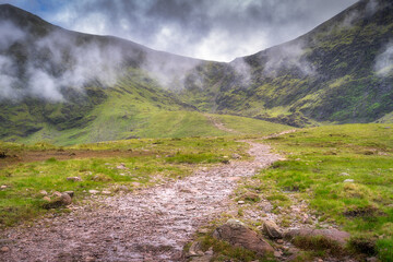 A serene view of lush green Carrauntoohil mountain and a winding path into the misty distance, ideal for nature lovers seeking beauty and adventure while traveling, Kerry, Ireland