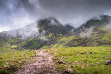 Enjoy a stunning scenic view featuring misty Carrauntoohil mountain and a winding pathway, beautifully surrounded by vibrant green grass and dramatic, fluffy clouds above, Kerry, Ireland