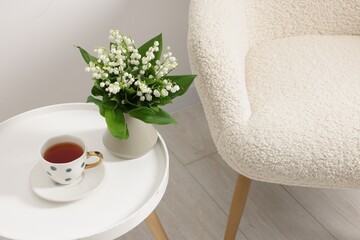 Bouquet of beautiful lily-of-the-valley in vase and cup of tea on coffee table near armchair indoors