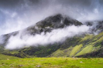 A breathtaking view of a majestic Carrauntoohil mountain elegantly shrouded in fluffy clouds, surrounded by vibrant green hills that stretch under a dramatic, colorful sky, Kerry, Ireland