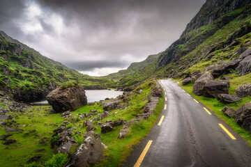 A captivating road meanders through lush, green hills and rugged rocky terrain of Gap of Dunloe, beneath dramatic, moody skies, creating the perfect backdrop for landscape photography, Kerry, Ireland