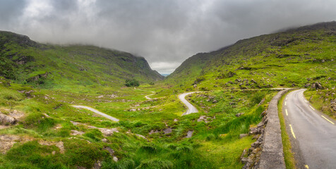 A peaceful and tranquil view of a winding road that gracefully traverses through a lush green Gap of Dunloe valley, beautifully framed by majestic mountain slopes beneath a cloudy sky, Kerry, Ireland