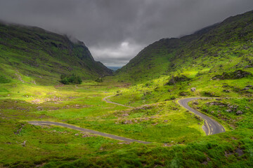 A winding road elegantly weaves its way through a vibrant green Gap of Dunloe valley, surrounded by majestic hills and moody clouds, creating a truly serene and picturesque landscape, Kerry, Ireland