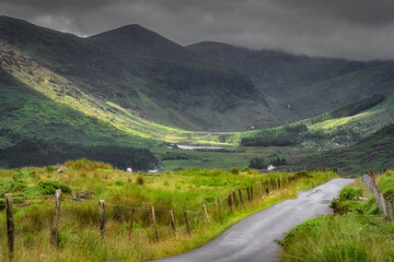 Discover and explore a breathtaking view of the rolling mountains and a tranquil Black Valley, beautifully illuminated by patches of warm sunlight that peek through the clouds, Kerry, Ireland