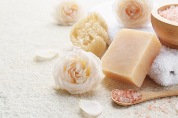 Soap bar, sea salt, loofah sponge, towel and flowers on white textured table, closeup. Space for text