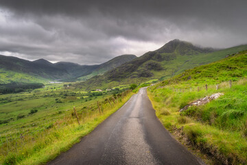 Explore a serene rural pathway that gracefully winds through vibrant, colorful hills of Black Valley, with majestic clouds encouraging the breathtaking views of the stunning landscape, Kerry, Ireland