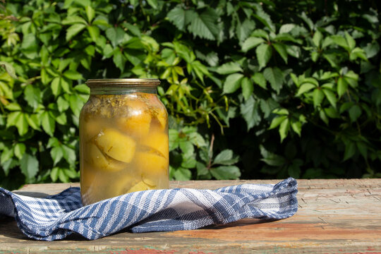 Canned homemade green tomatoes with herbs in a glass jar on a wooden table against a background of greenery on a sunny summer day. Copy space. 
