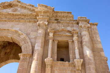 Fototapeta premium Visitors admire the intricate design of the Arch of Hadrian, a monumental structure in Jerash, Jordan. The historic site showcases ancient Roman architecture under a clear blue sky.