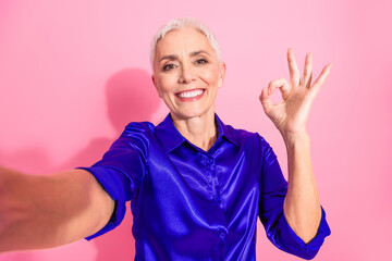 Confident mature woman in stylish blue shirt showing ok gesture on pink background promoting...