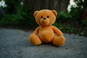 A soft orange teddy bear is positioned on the ground with a backdrop of lush greenery The warm late...