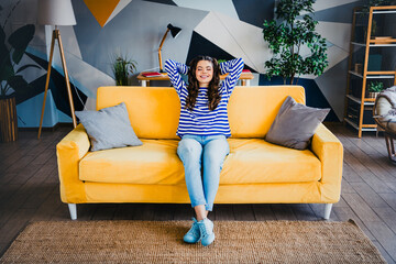 Cheerful young woman relaxing on a bright yellow sofa in a modern living room with a cozy and...