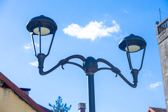 An elegant vintage street lamp with two ornate light fixtures stands against a clear blue sky. a glimpse of traditional rooftops adds a nostalgic touch. perfect for themes of nostalgia and urban