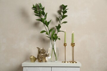 Elegant candlesticks with burning candles, branches in vase and other decor on dresser near beige...