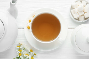Delicious chamomile tea in cup, fresh flowers, sugar cubes and teapot on white wooden table, flat lay