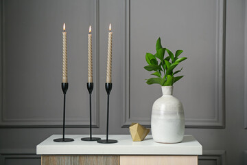 Elegant candlesticks with burning candles, branches in vase and other decor on dresser near grey...