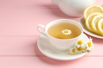 Delicious chamomile tea in cup, fresh flowers and lemon on pink wooden table, closeup. Space for text