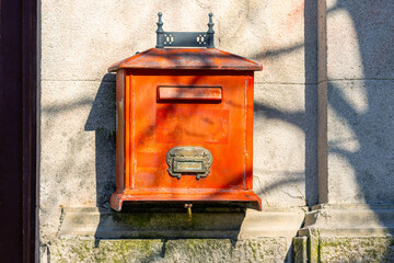 An ornate vintage red mailbox adorns a textured stone wall. shadows play across the surface, creating a dynamic contrast between light and color, enhancing its classic appeal.