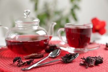 Delicious hibiscus tea in glass cup, teapot, dry roselle sepals and spoons on red mat, closeup