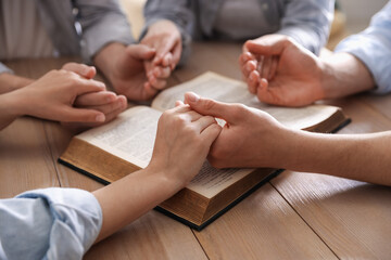 Family with Bible praying at wooden table indoors, closeup