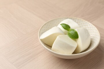 Pieces of delicious mozzarella cheese and basil in bowl on wooden table, closeup. Space for text