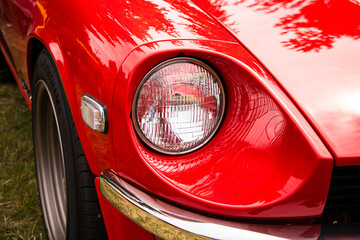 Detailed view of the headlight on a bright red sports car. The chrome and glass reflect light, with grass and other vintage cars blurred in the background
