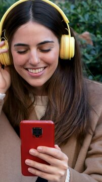 Happy Young Woman Listening to Music with Headphones and Using Smartphone Outdoors