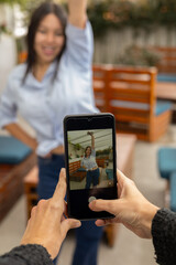 A vibrant young woman smiles warmly as a smartphone camera focuses on her outdoors, blending personal expression with everyday technology in a natural setting.

