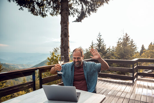 Smiling middle aged man enjoying virtual meeting with wine on a mountain deck